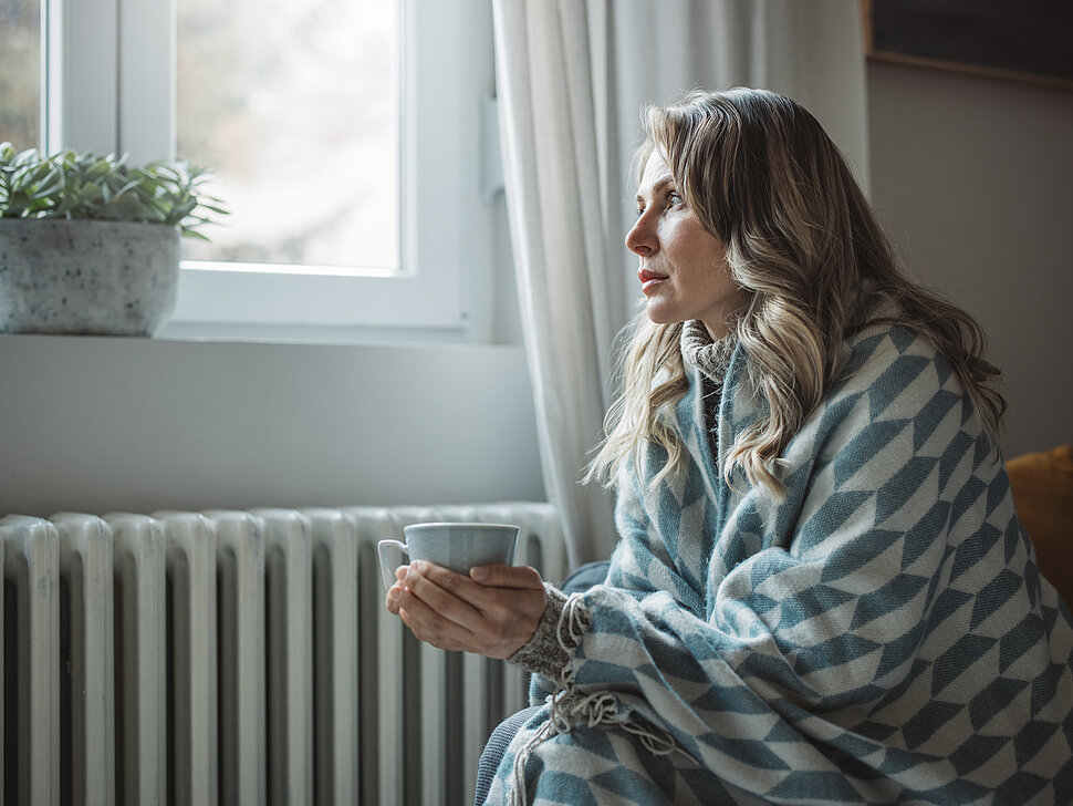  Frau in Decke und mit Tasse sitzt vor der Heizung