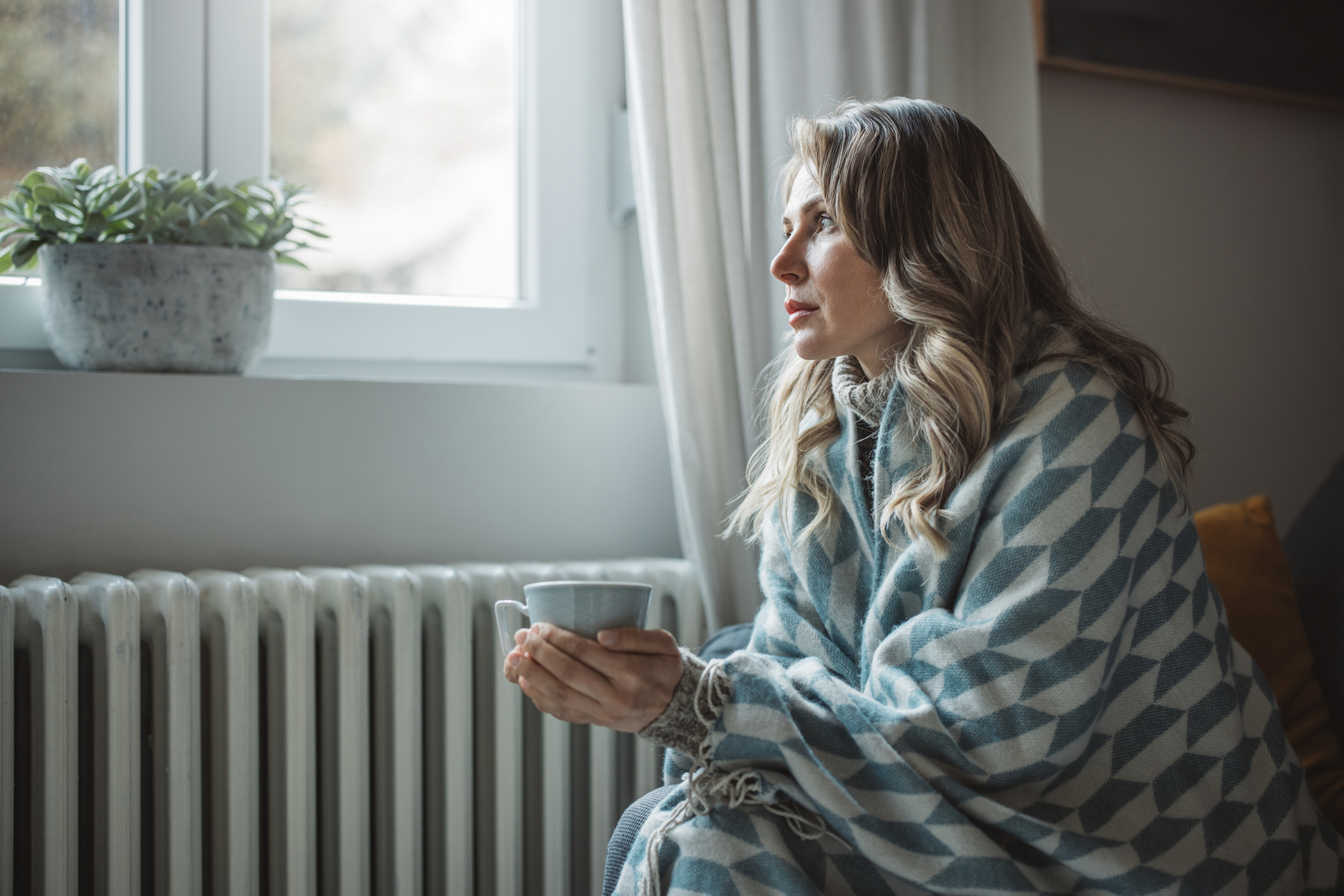  Frau in Decke und mit Tasse sitzt vor der Heizung