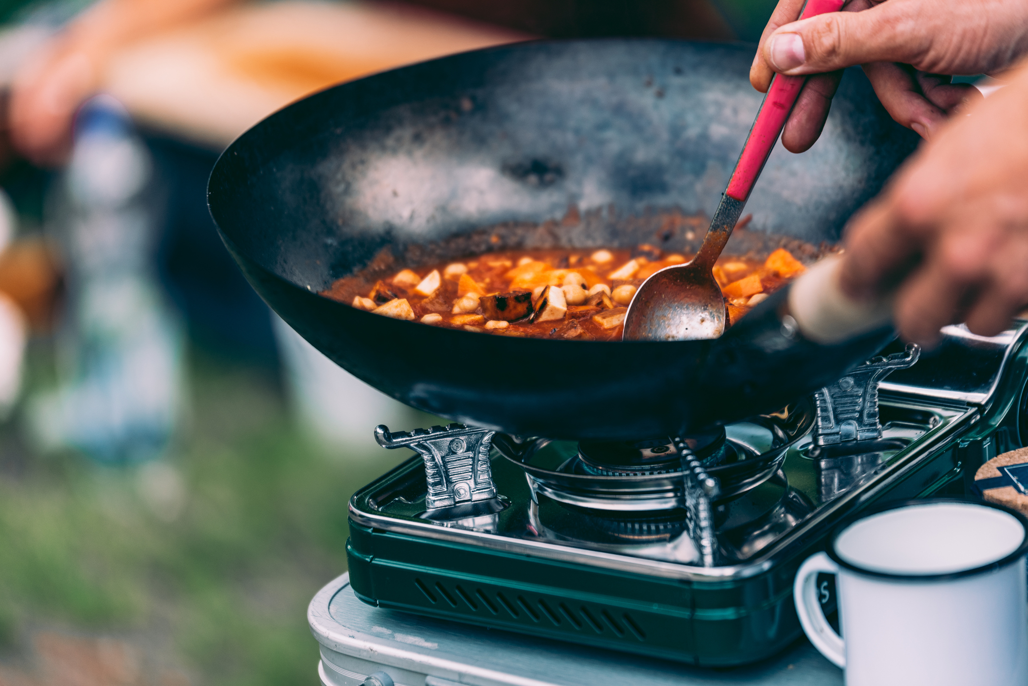 Hand rührt ein Gericht in einer Wok-Pfanne auf einem Camping-Herd